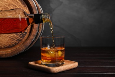 Photo of Pouring whiskey from bottle into glass with ice cubes near wooden barrel on table, space for text