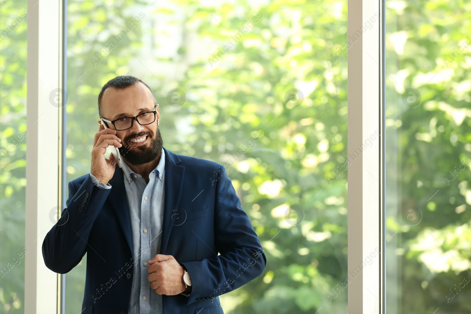 Photo of Portrait of handsome mature man in elegant suit talking on mobile phone near window. Space for text