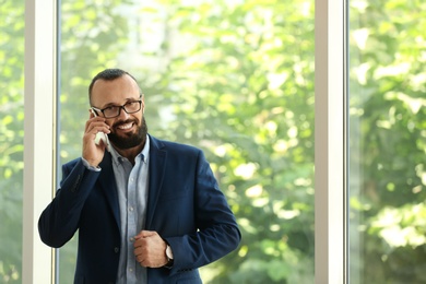 Photo of Portrait of handsome mature man in elegant suit talking on mobile phone near window. Space for text