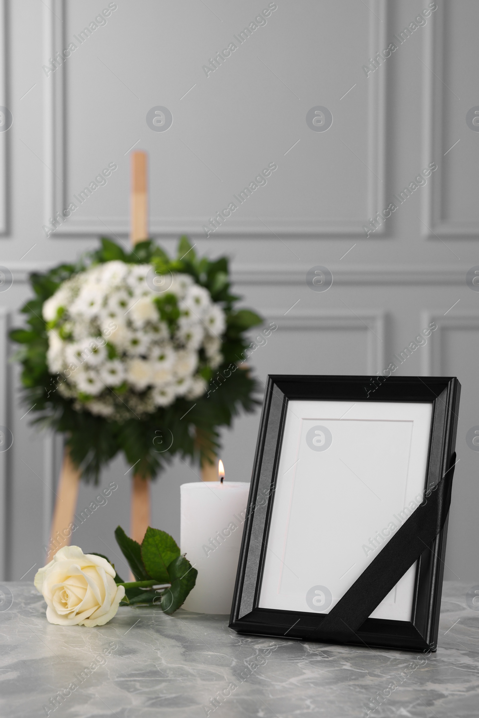 Photo of Photo frame with black ribbon, rose, burning candle on light grey table and wreath of flowers near wall indoors. Funeral attributes
