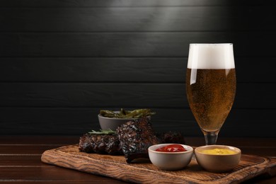 Photo of Glass of beer, delicious grilled ribs and sauces on wooden table. Space for text