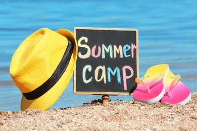 Photo of Little blackboard with text SUMMER CAMP, hat and flip flops on sand near sea