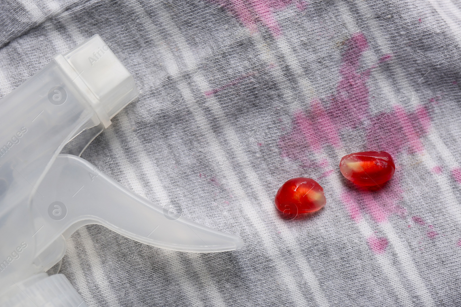 Photo of Striped shirt with fruit juicy stains, detergent and pomegranate seeds as background, closeup