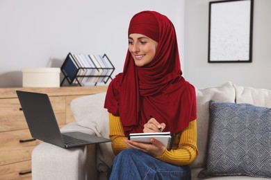 Muslim woman writing notes near laptop at couch in room