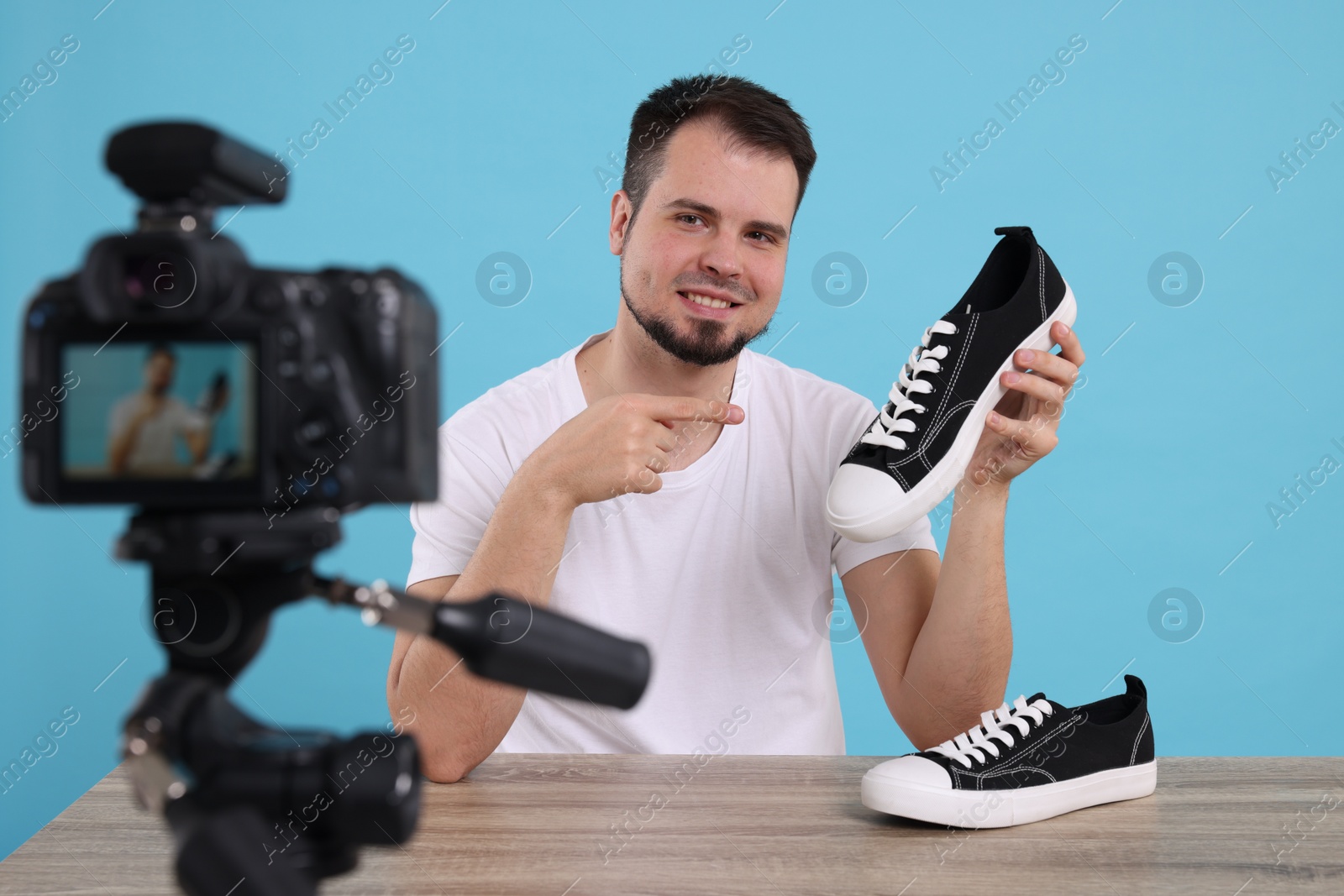 Photo of Smiling fashion blogger showing sneakers while recording video at table against light blue background