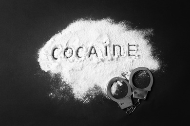 Photo of Heap of white powder with written word Cocaine and handcuffs on black background, top view