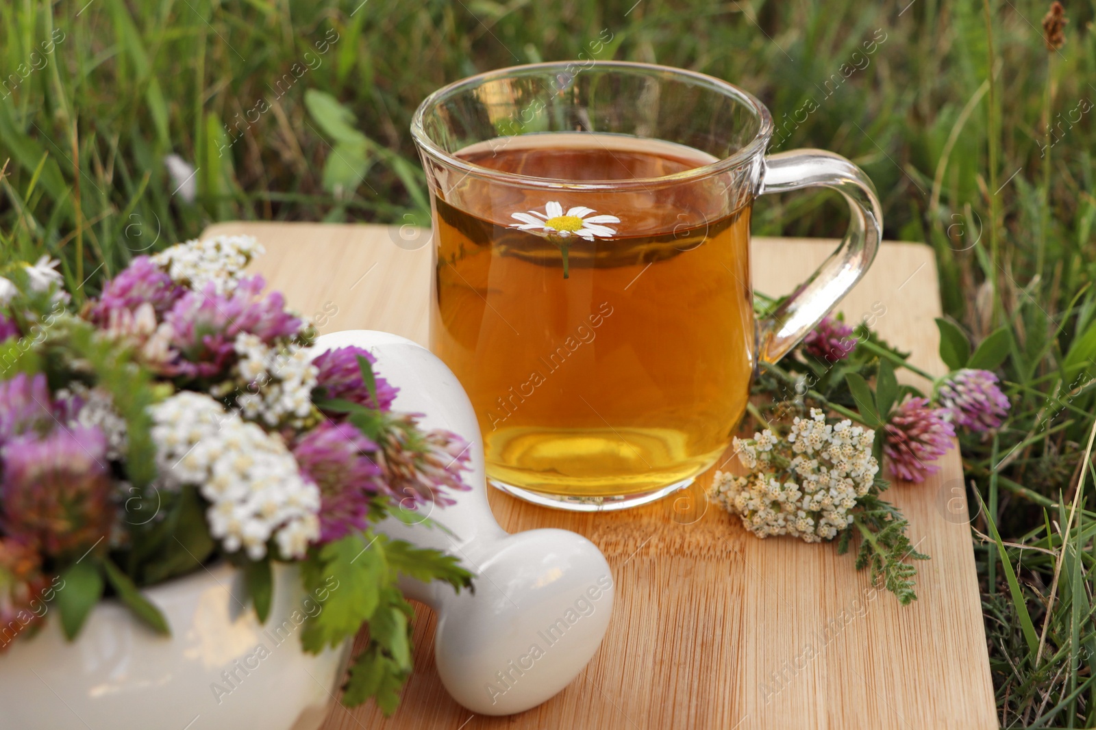 Photo of Cup of aromatic herbal tea, pestle and ceramic mortar with different wildflowers on green grass outdoors