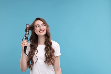 Beautiful young woman using curling hair iron on light blue background, space for text