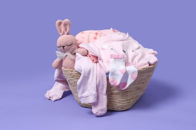 Laundry basket with baby clothes and soft toy on light purple background