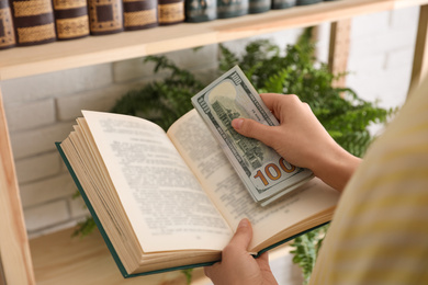 Woman hiding dollar banknotes in book indoors, closeup. Money savings