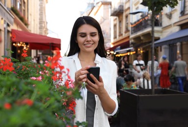 Photo of Young woman with smartphone near beautiful flowers on city street