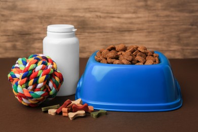 Photo of Bowl with dry pet food, bottle of vitamins and toy on brown surface