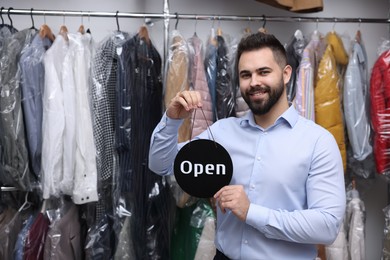 Photo of Dry-cleaning service. Happy worker holding Open sign near clothes in plastic bags indoors