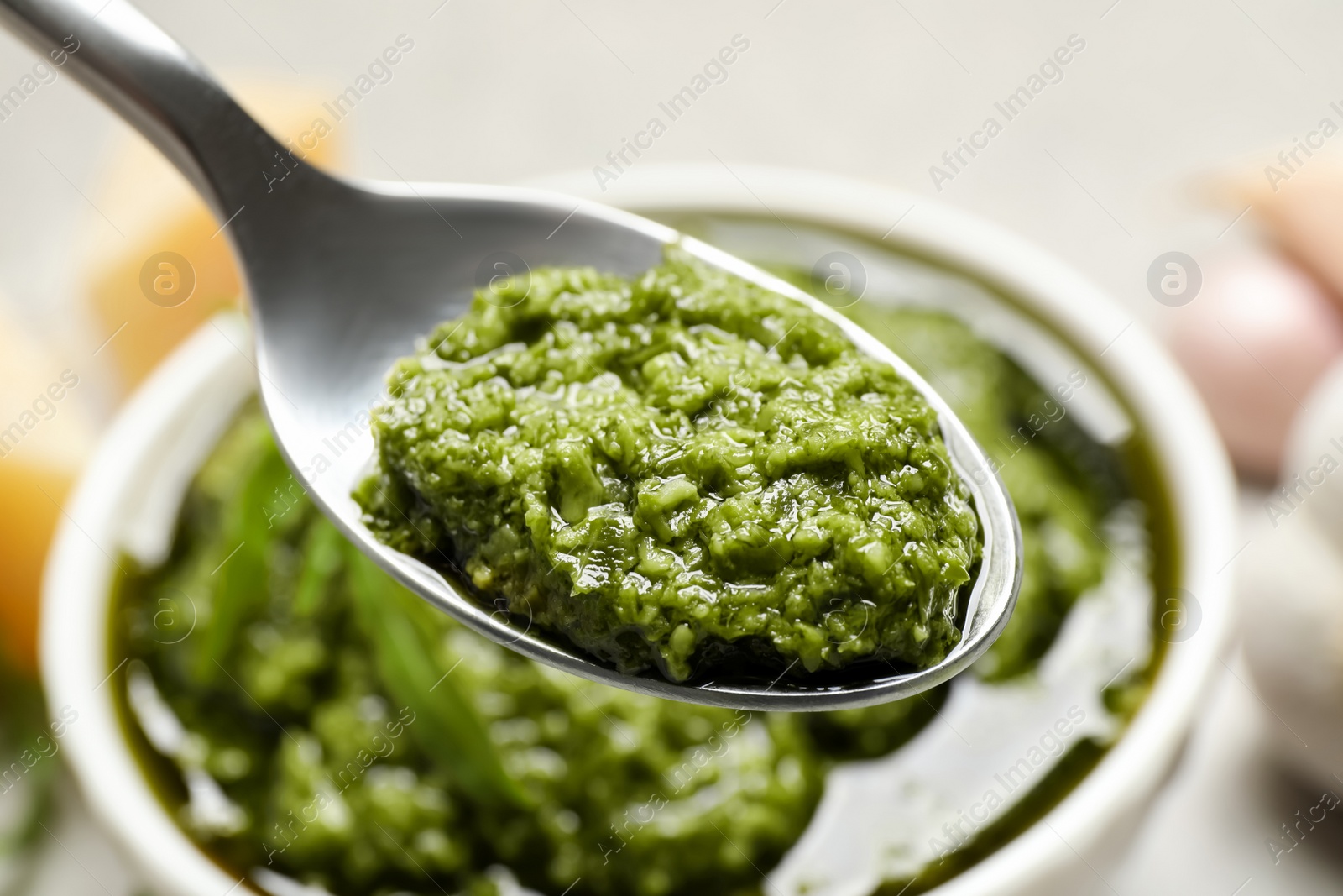 Photo of Spoon of tasty arugula pesto near bowl with sauce, closeup