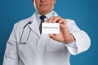 Photo of Doctor holding business card with word HEMORRHOID on blue background, closeup