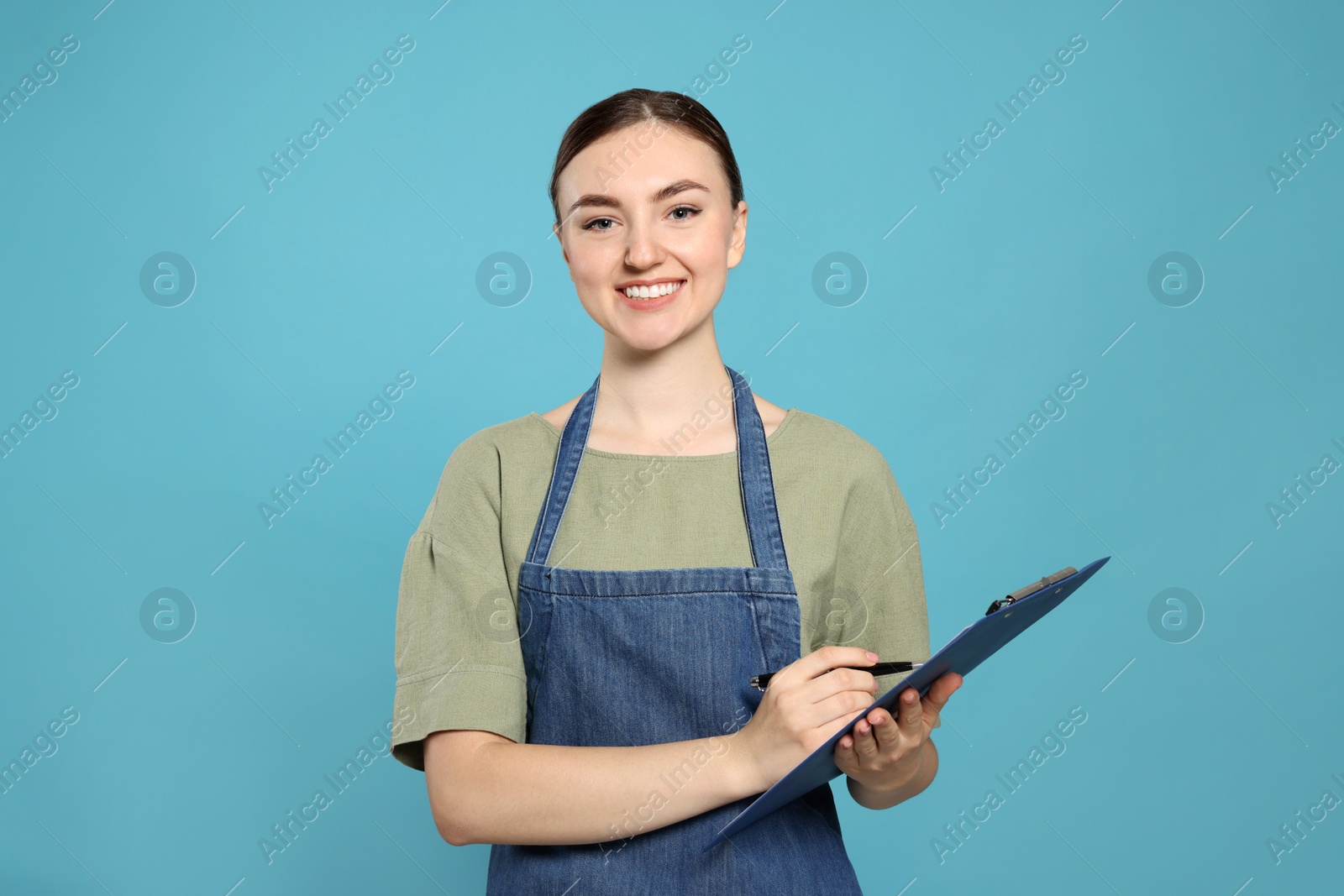 Photo of Beautiful young woman in clean denim apron with clipboard on light blue background