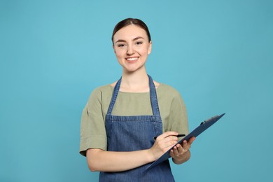 Beautiful young woman in clean denim apron with clipboard on light blue background