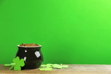 Photo of Pot of gold coins and clover leaves on wooden table against green background, space for text. St. Patrick's Day celebration