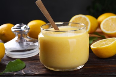 Photo of Delicious lemon curd in glass jar, fresh citrus fruits and spoon on wooden table