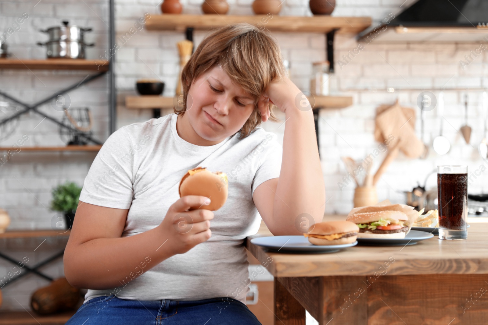 Photo of Emotional overweight boy at table with fast food in kitchen