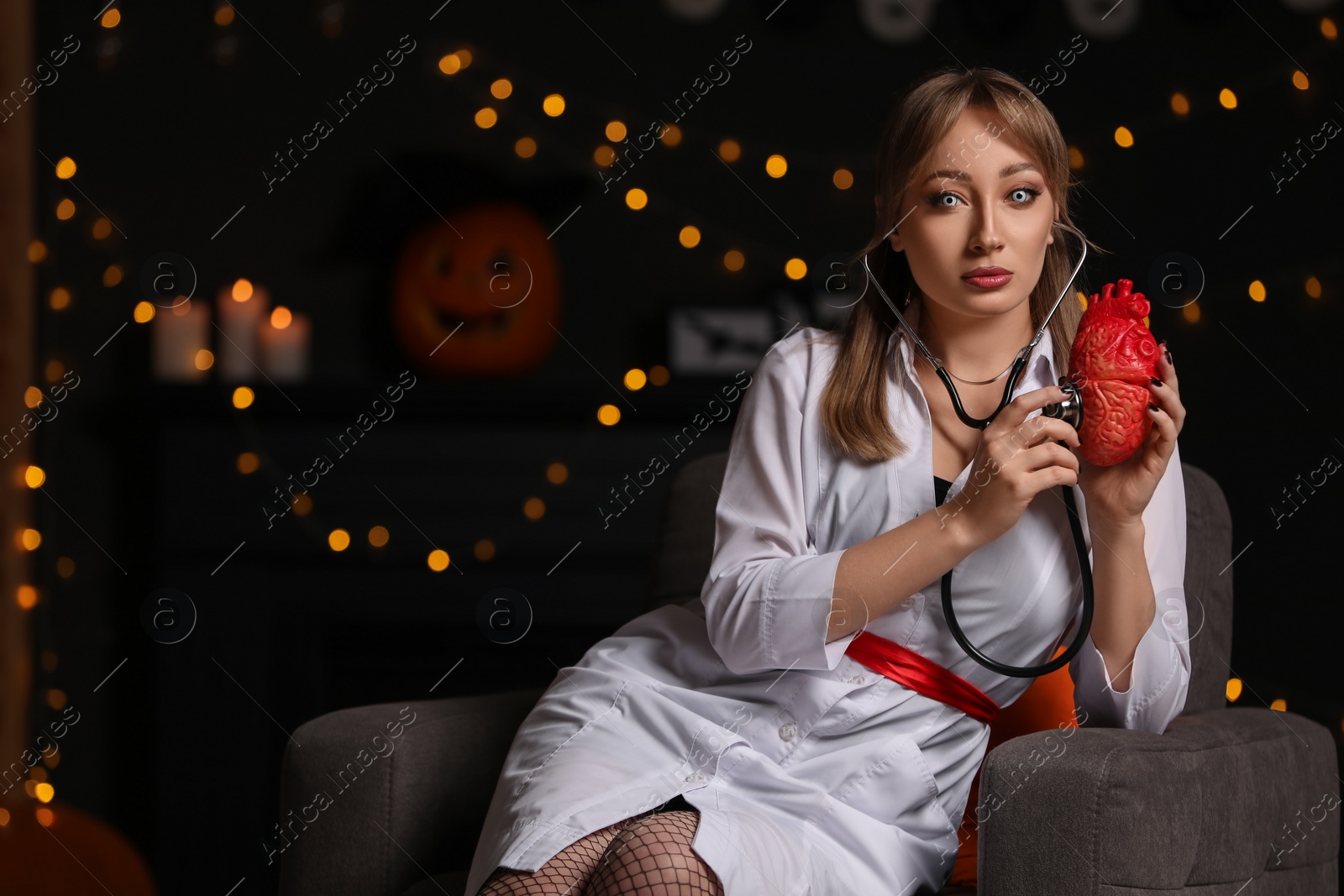 Photo of Woman in scary nurse costume with heart model and stethoscope against blurred lights indoors, space for text. Halloween celebration