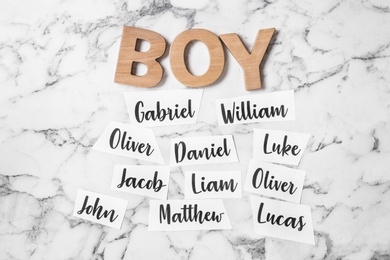 Paper sheets with different baby names and wooden letters on white marble background, flat lay