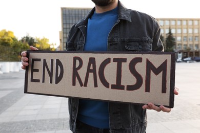 Photo of Young man holding sign with phrase End Racism outdoors, closeup
