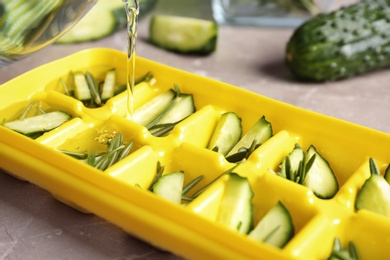 Photo of Pouring water into ice cube tray with cucumber and rosemary on table, closeup