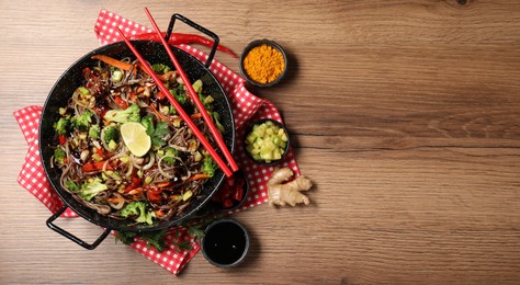 Photo of Stir-fry. Tasty noodles with meat in wok, chopsticks and ingredients on wooden table, flat lay. Space for text