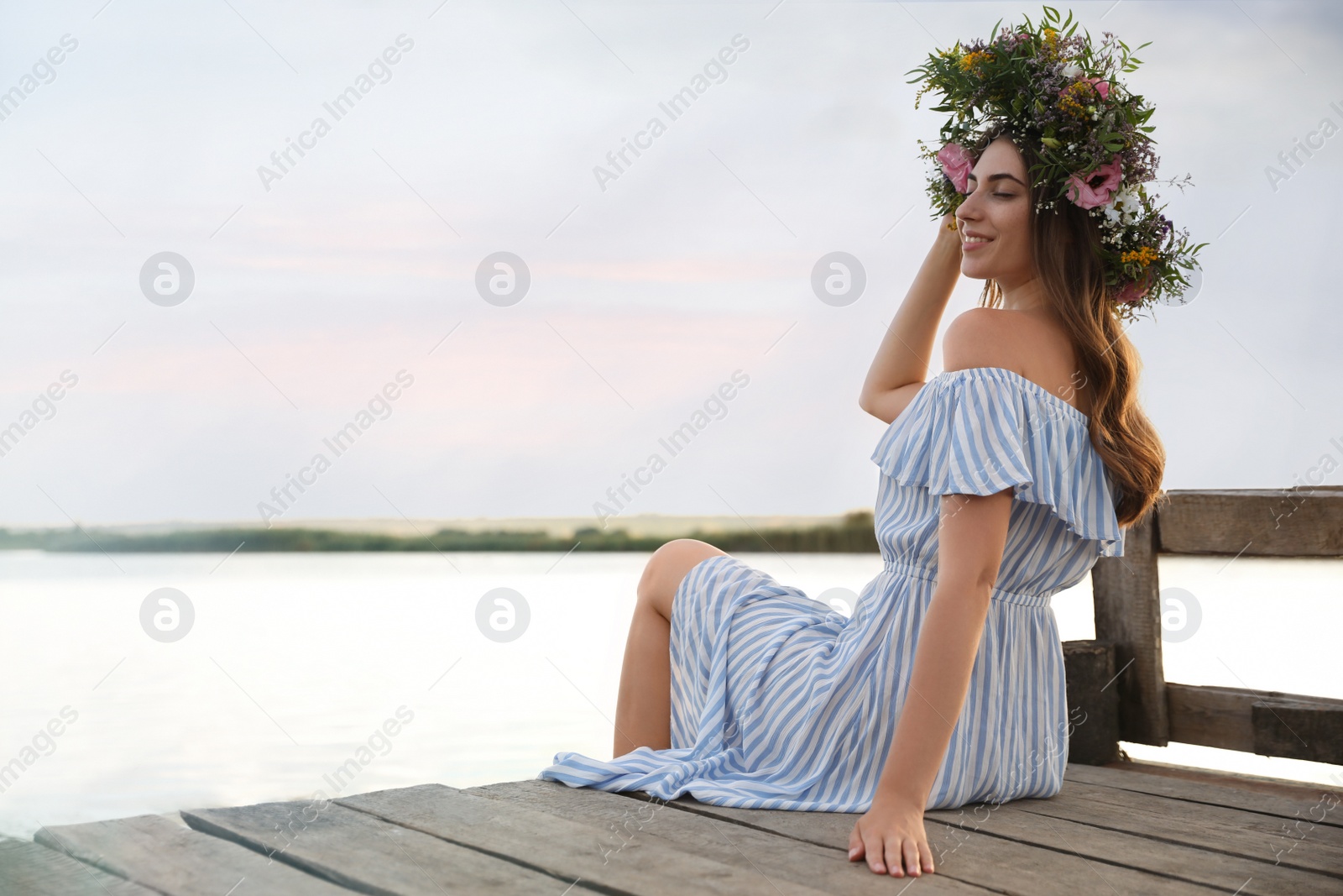 Photo of Young woman wearing wreath made of beautiful flowers on pier near river