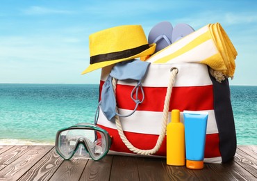 Image of Stylish bag with beach accessories on wooden surface near seashore