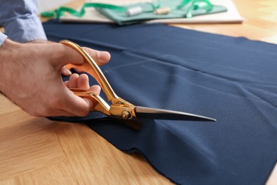 Photo of Man cutting blue fabric with scissors at wooden table, closeup
