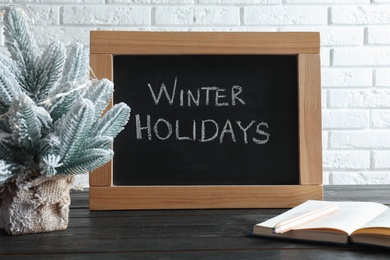 Photo of Chalkboard with phrase Winter Holidays, small Christmas tree and book on black wooden table near white brick wall