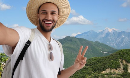 Image of Smiling young man in straw hat taking selfie and showing peace sign in mountains