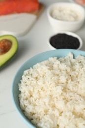 Photo of Cooked rice in bowl and other ingredients for sushi on white table, closeup