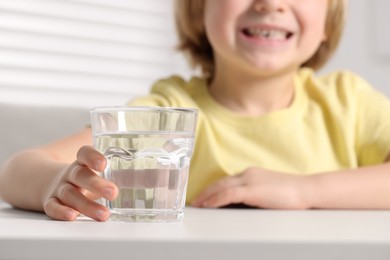 Photo of Little boy holding glass of fresh water at white table indoors, closeup