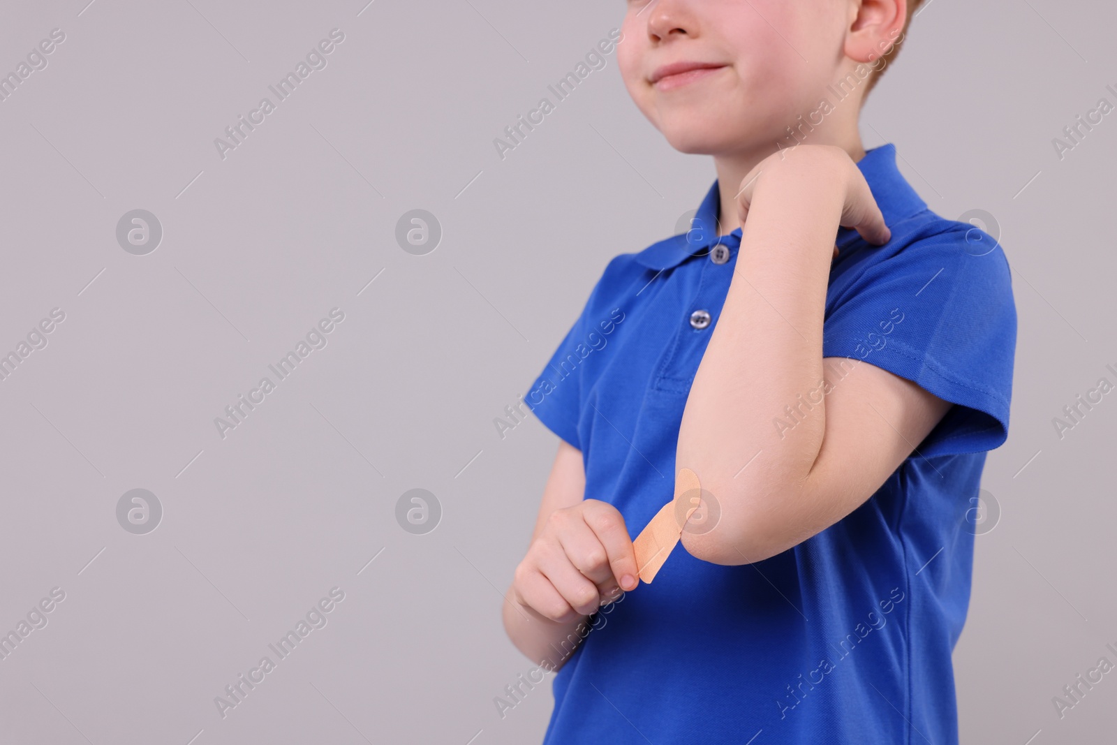 Photo of Little boy putting sticking plaster onto elbow against light grey background, closeup. Space for text