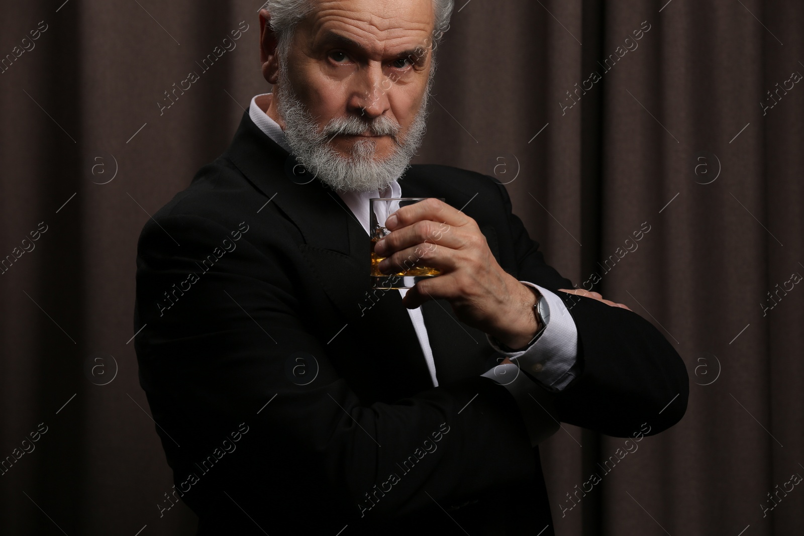 Photo of Senior man in formal suit holding glass of whiskey with ice cubes on brown background