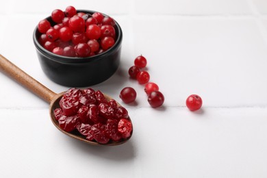 Tasty dried cranberries in spoon and fresh ones on white tiled table. Space for text