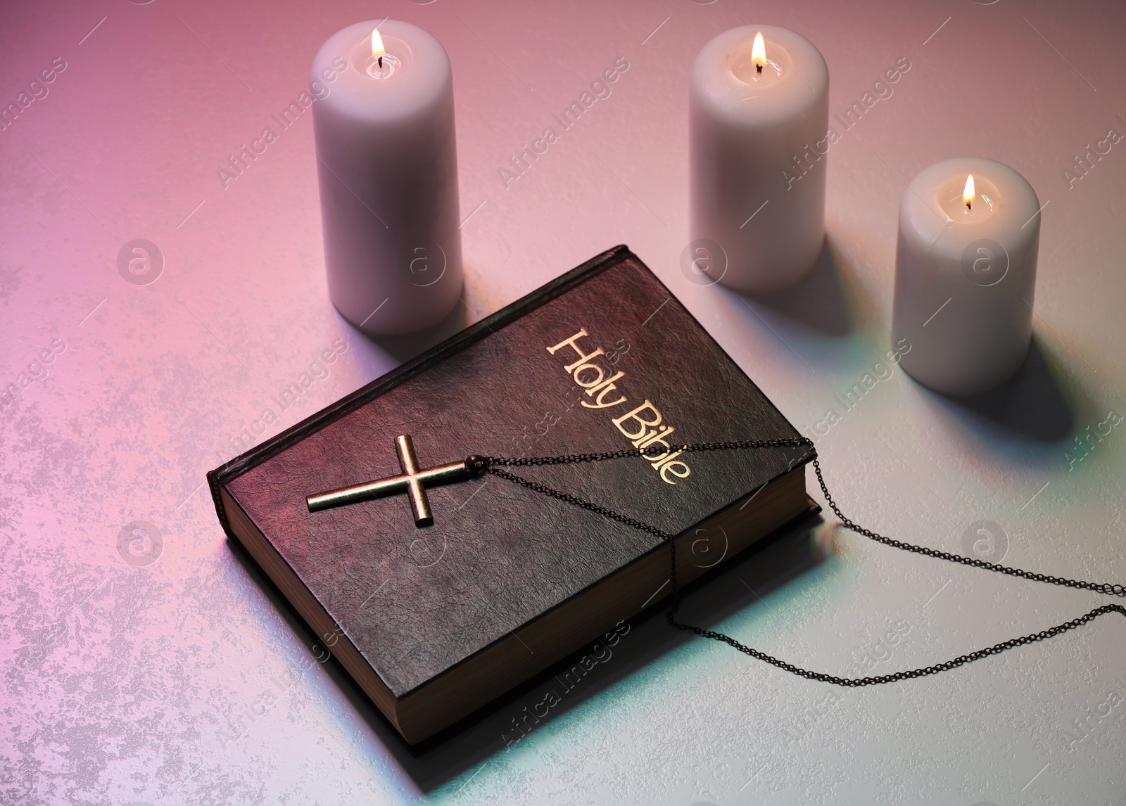 Photo of Cross, burning candles and Bible on textured table in color lights, above view. Religion of Christianity