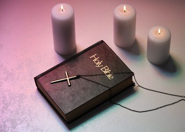 Cross, burning candles and Bible on textured table in color lights, above view. Religion of Christianity