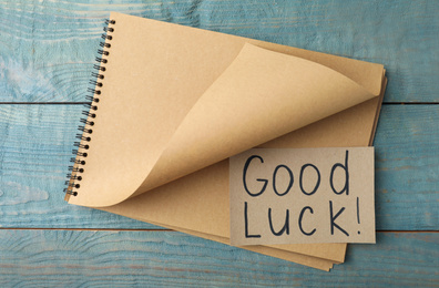 Photo of Sheet of paper with phrase GOOD LUCK and notebook on light blue wooden table, top view