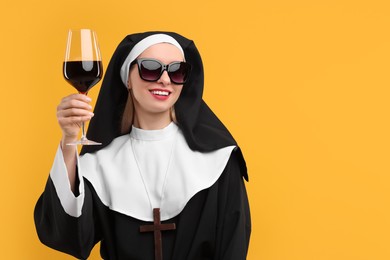 Photo of Happy woman in nun habit and sunglasses holding glass of wine against orange background. Space for text