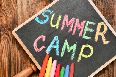 Photo of Little blackboard with text SUMMER CAMP chalked in different colors on table, top view