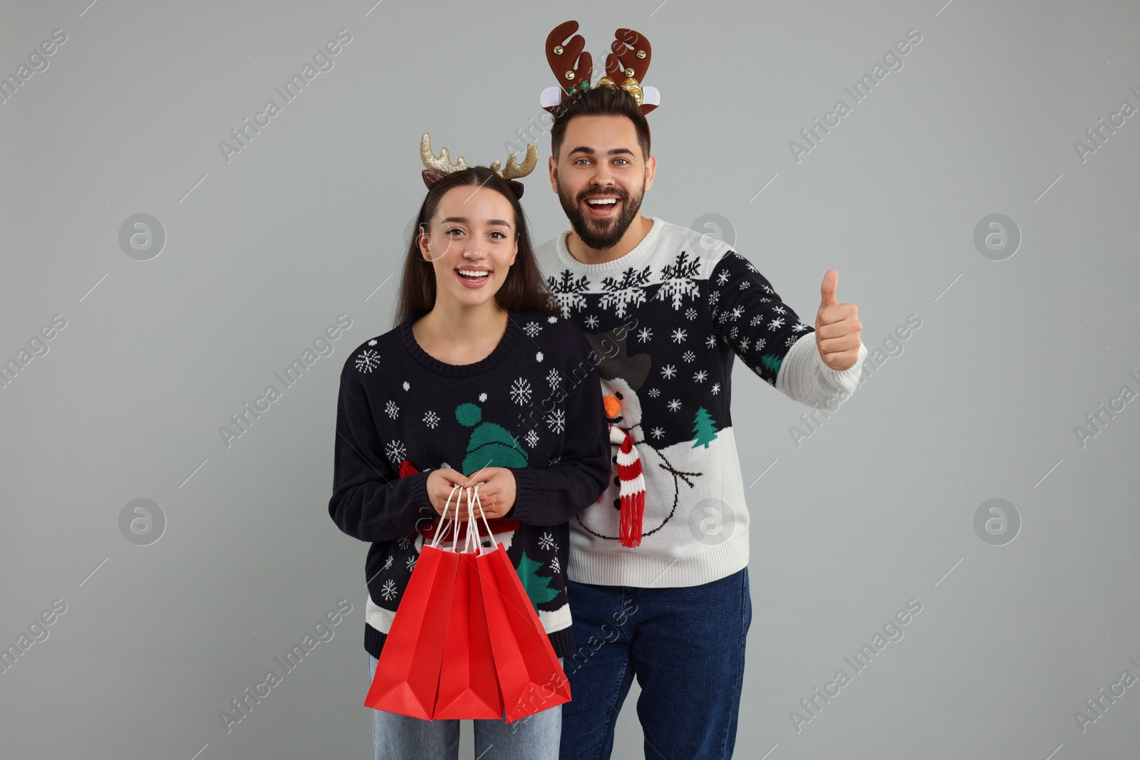 Photo of Happy young couple in Christmas sweaters and reindeer headbands with shopping bags on grey background