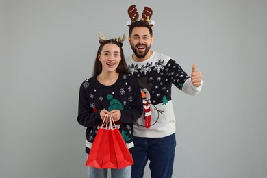 Photo of Happy young couple in Christmas sweaters and reindeer headbands with shopping bags on grey background