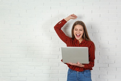 Emotional young woman with laptop celebrating victory near brick wall. Space for text