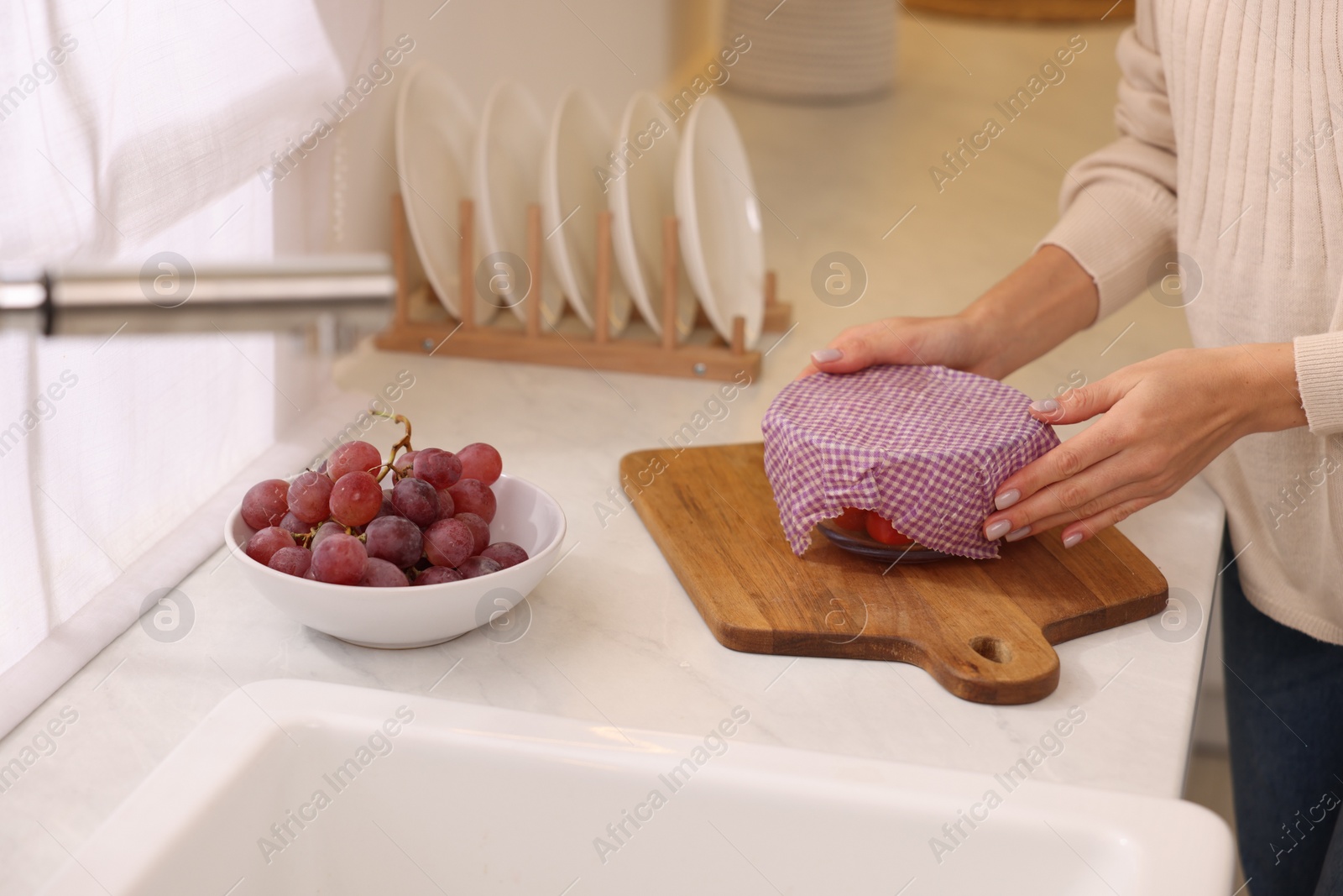 Photo of Woman packing bowl of fresh tomatoes into beeswax food wrap at white countertop in kitchen, closeup