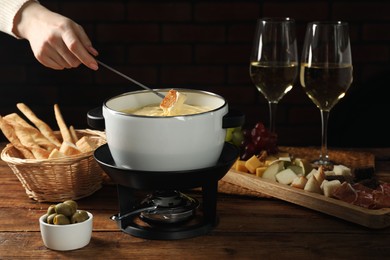 Photo of Woman dipping piece of bread into fondue pot with melted cheese at wooden table, closeup