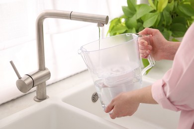 Photo of Woman filling filter jug with water from tap in kitchen, closeup